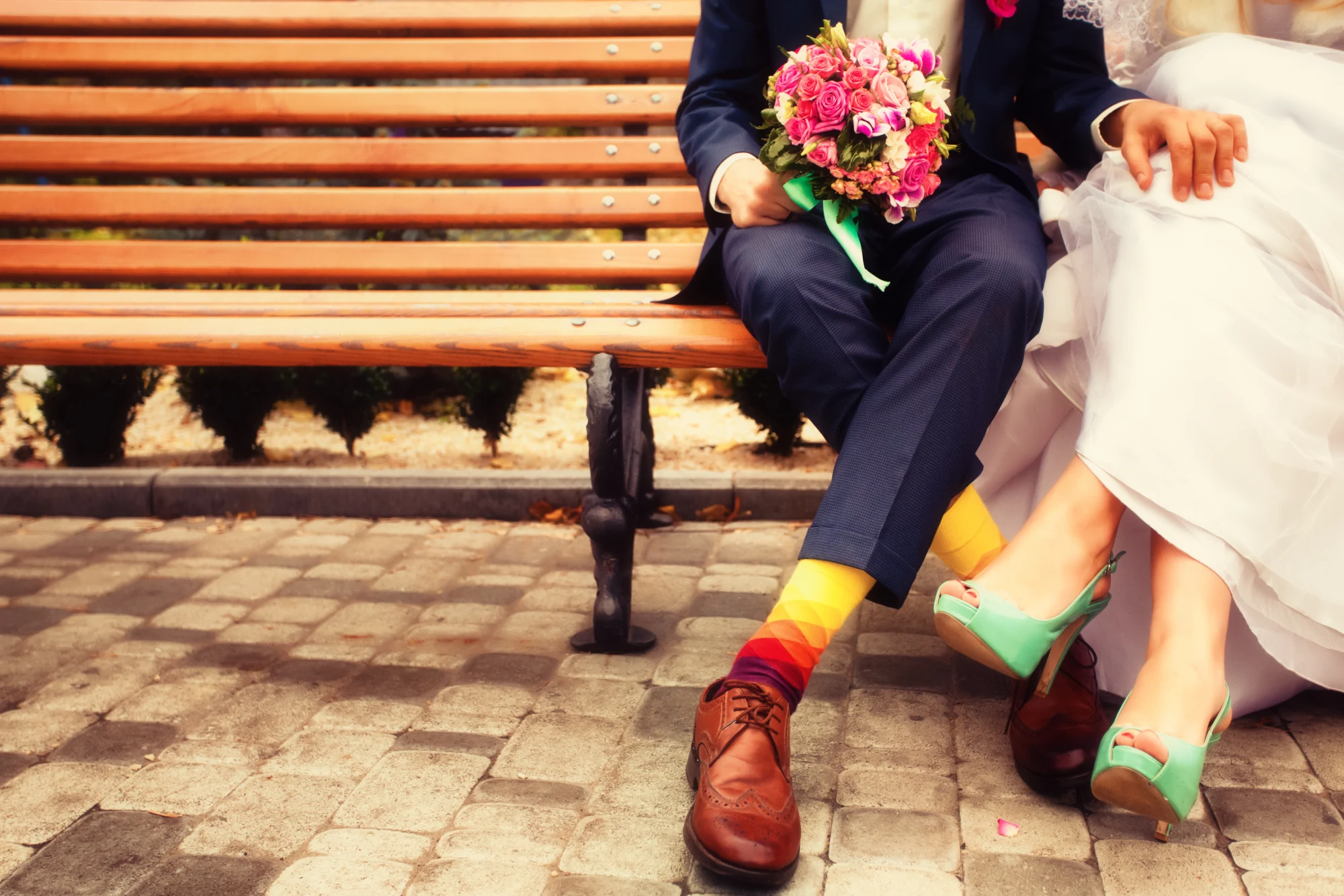 A bride and groom seated on a bench with a colorful rose bouquet and bright wedding heels